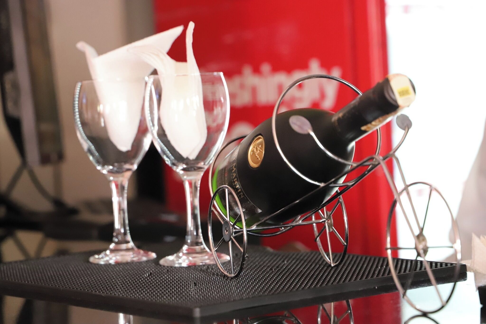 A bottle of champagne on a stand with two glasses filled with a white liquid, likely champagne, in a setting with a red backdrop.