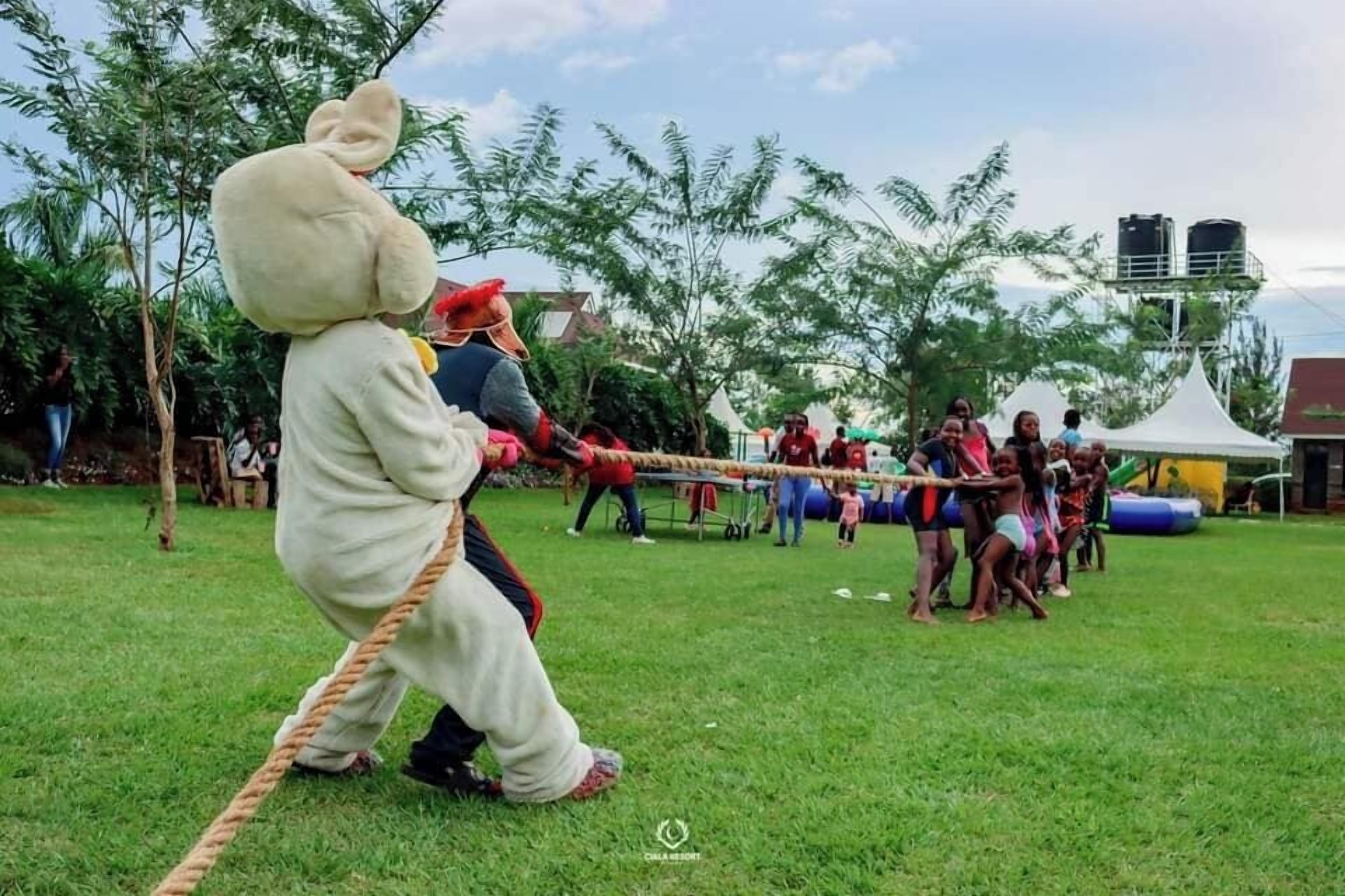 Un groupe de personnes joue à un jeu de tir à la corde dans un champ herbeux, avec des arbres et des tentes en arrière-plan.