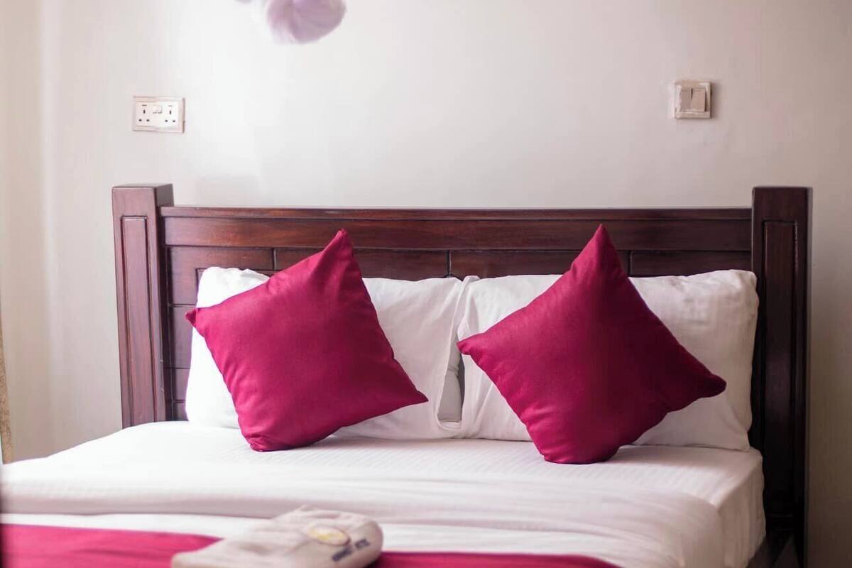 A neatly made bed with white linens and two bright pink pillows, a wooden headboard, and a white bedside table with a folded white towel.