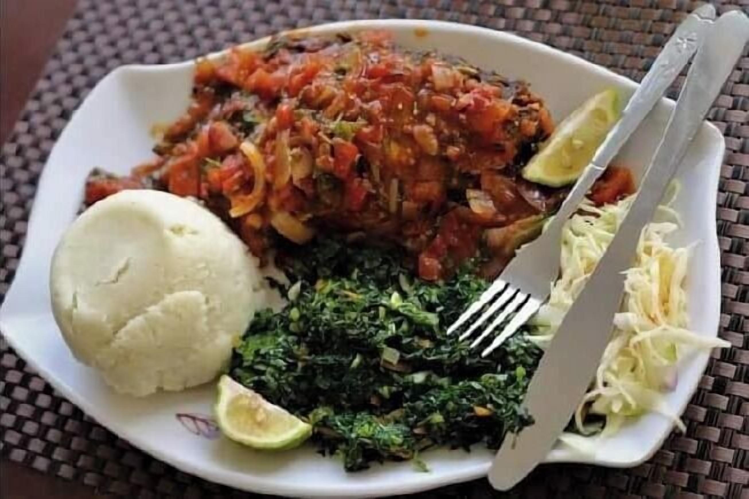 A plate with a variety of food including a mound of white rice, a green leafy vegetable, a tomato-based dish, and a side of shredded cabbage, accompanied by a fork and knife.