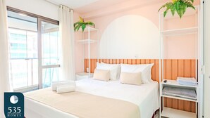 A hotel room with a large bed, a wooden headboard, a white shelf with a plant, and a view of the city through the window.