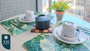 A table with a tropical leaf-patterned placemat, a black pot, two white cups, and a potted plant with yellow flowers.