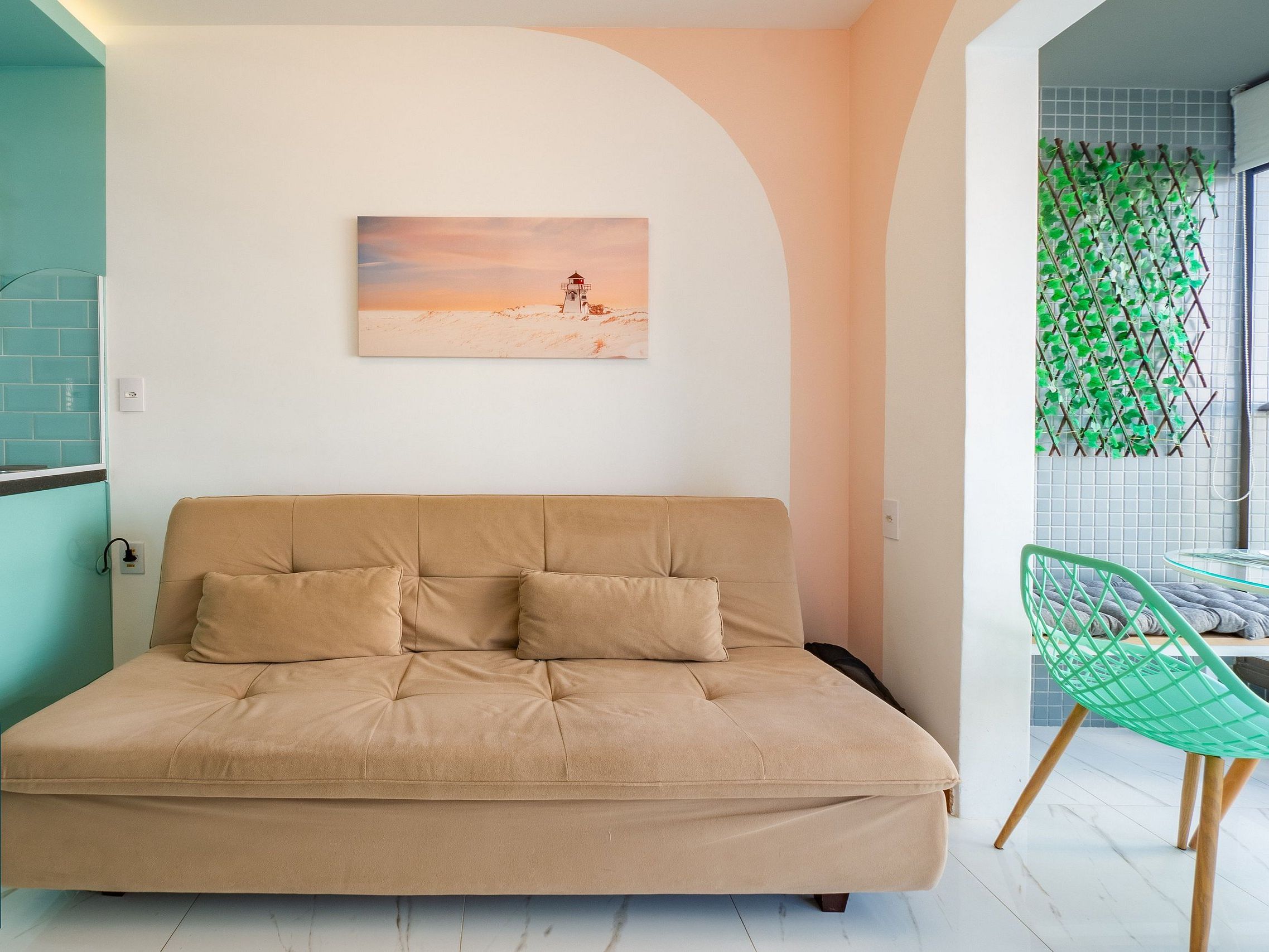 A modern living room with a beige sofa, a green chair, and a painting of a lighthouse on the wall.