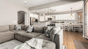 A modern living room with a sectional sofa, a dining area with a wooden table and chairs, and a kitchen with white cabinets and stainless steel appliances.