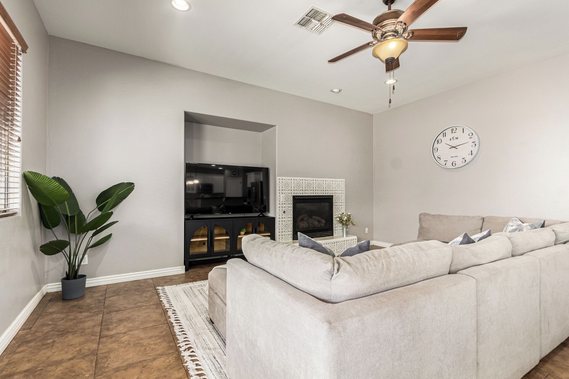A living room with a beige sofa, a fireplace, a ceiling fan, and a wall clock.