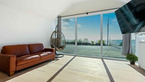 Une pièce spacieuse dotée d’une grande porte-fenêtre coulissante donnant sur un espace extérieur, d’un canapé en cuir marron, d’un fauteuil suspendu et d’un téléviseur à écran plat fixé au mur.