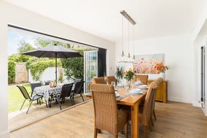 Een eetgedeelte met een grote tafel, stoelen en een parasol. Achter de glazen deuren is een tuin zichtbaar.