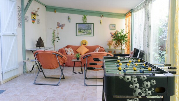 Een kamer met een tafelvoetbaltafel, oranje stoelen, een bank, een kleine tafel en een raam met gordijnen.