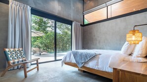 Une chambre moderne avec un grand lit, une table de chevet en bois, une chaise avec un coussin aux motifs géométriques et une vue sur la verdure à l’extérieur.