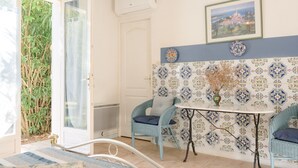 A room with a white door, a window with a view of greenery, a wall with patterned tiles, a table with a vase, and two blue chairs.
