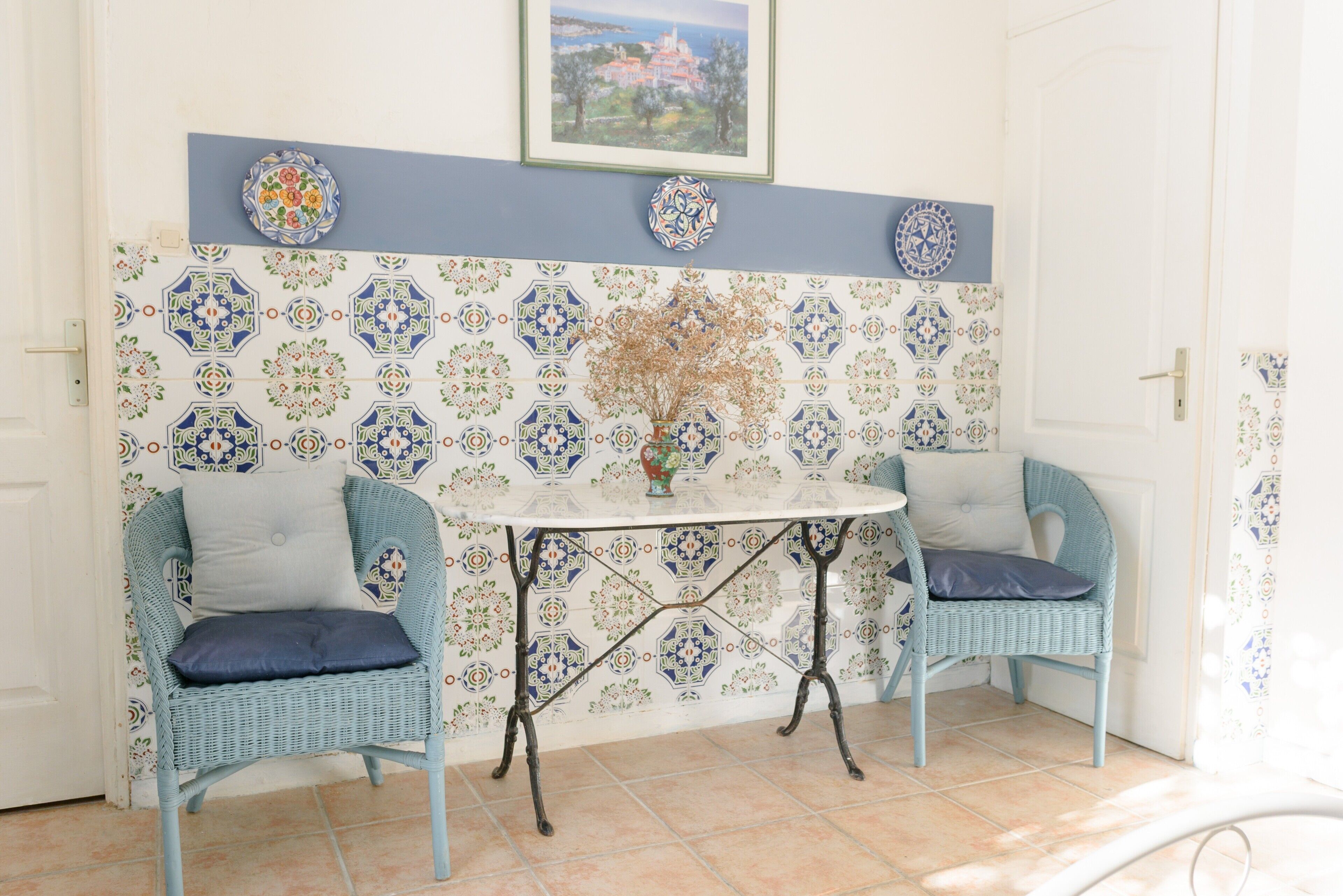 Une pièce d’angle avec des carreaux décoratifs, une table surmontée d’un vase, deux chaises en osier et une porte.