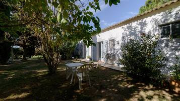 A white building with a tiled roof, a patio with a table and chairs, and a garden area with trees and shrubs.