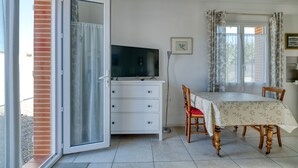 A room with a sliding glass door, a white dresser, a television, a dining table with chairs, and a window with curtains.