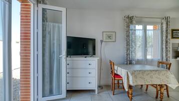 Una stanza con una porta scorrevole in vetro, una cassettiera bianca, un televisore, un tavolo da pranzo con sedie e una finestra con tende.