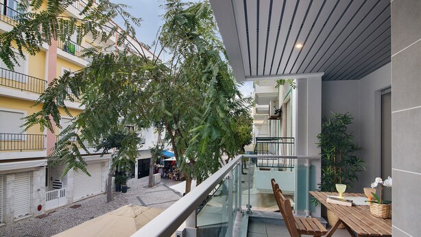 A balcony with a wooden table and chairs, a glass railing, and a view of a courtyard with trees and buildings. - Apartment 'Eanes 22 Beach House' with Balcony, Wi-Fi and Air Conditioning (Monte Gordo)