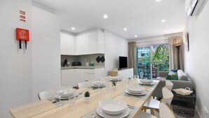 A modern dining area with a long table set for six, white cabinets, a kitchen area, and a view of greenery outside. - Apartment 'Eanes 22 Beach House' with Balcony, Wi-Fi and Air Conditioning (Monte Gordo)