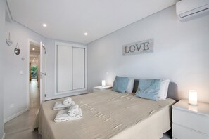 A bedroom with a bed, nightstands, a wall-mounted air conditioner, and a door leading to another room.