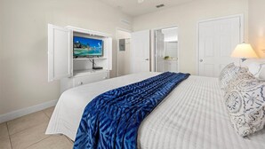 Une chambre à coucher avec un lit, un téléviseur posé sur un meuble, un ventilateur de plafond et une porte donnant sur une autre pièce.