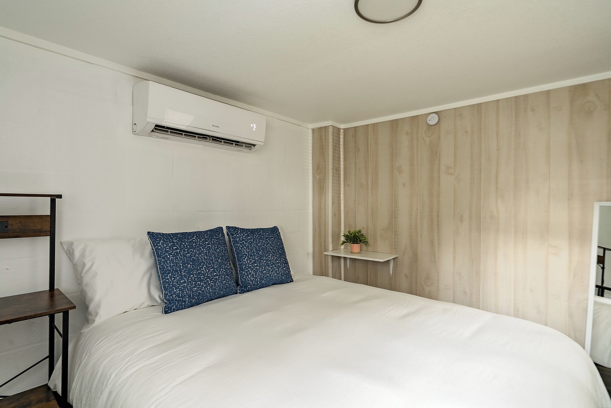 Een slaapkamer met een bed, wit beddengoed, een blauw gestreept kussen, een klein plankje met een plant en een wandairconditioner.
