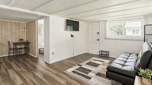Una sala de estar moderna con un sofá de cuero negro, una mesa de comedor de madera con sillas, un televisor de pantalla plana montado en la pared y un ventanal que proporciona luz natural.