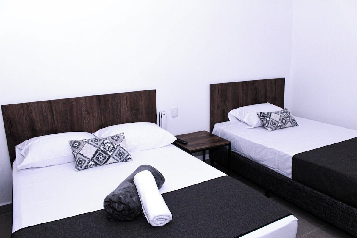 Une chambre d’hôtel avec deux lits simples, chacun doté d’une tête de lit en bois, de draps blancs et d’oreillers à motifs. Une petite table de chevet en bois est placée entre les lits, et une télécommande y est posée. Une serviette enroulée est posée sur le sol, devant les lits.