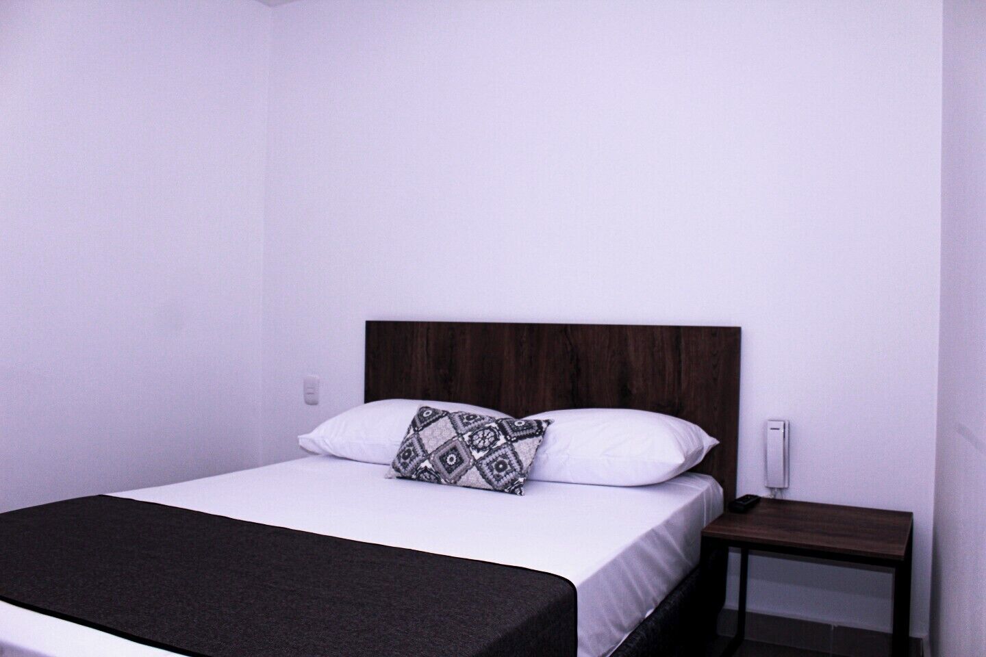 A single bed with a dark headboard, white bedding, and a patterned pillow. There is a small wooden nightstand beside the bed.