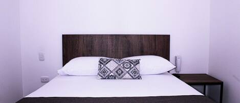 Uma cama com cabeceira escura, roupa de cama branca e almofada estampada.