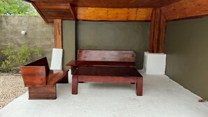Un banc en bois, un jardinier en métal et un bloc blanc placés sous une structure de toit en bois.