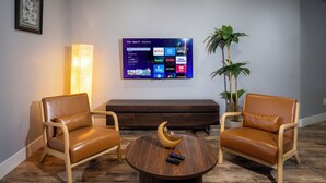 Ein Wohnzimmer mit zwei Ledersesseln, einem runden Couchtisch mit Mond- und Sterndekoration, einem Flachbildfernseher, der Streamingdienste zeigt, und einer Zimmerpflanze.