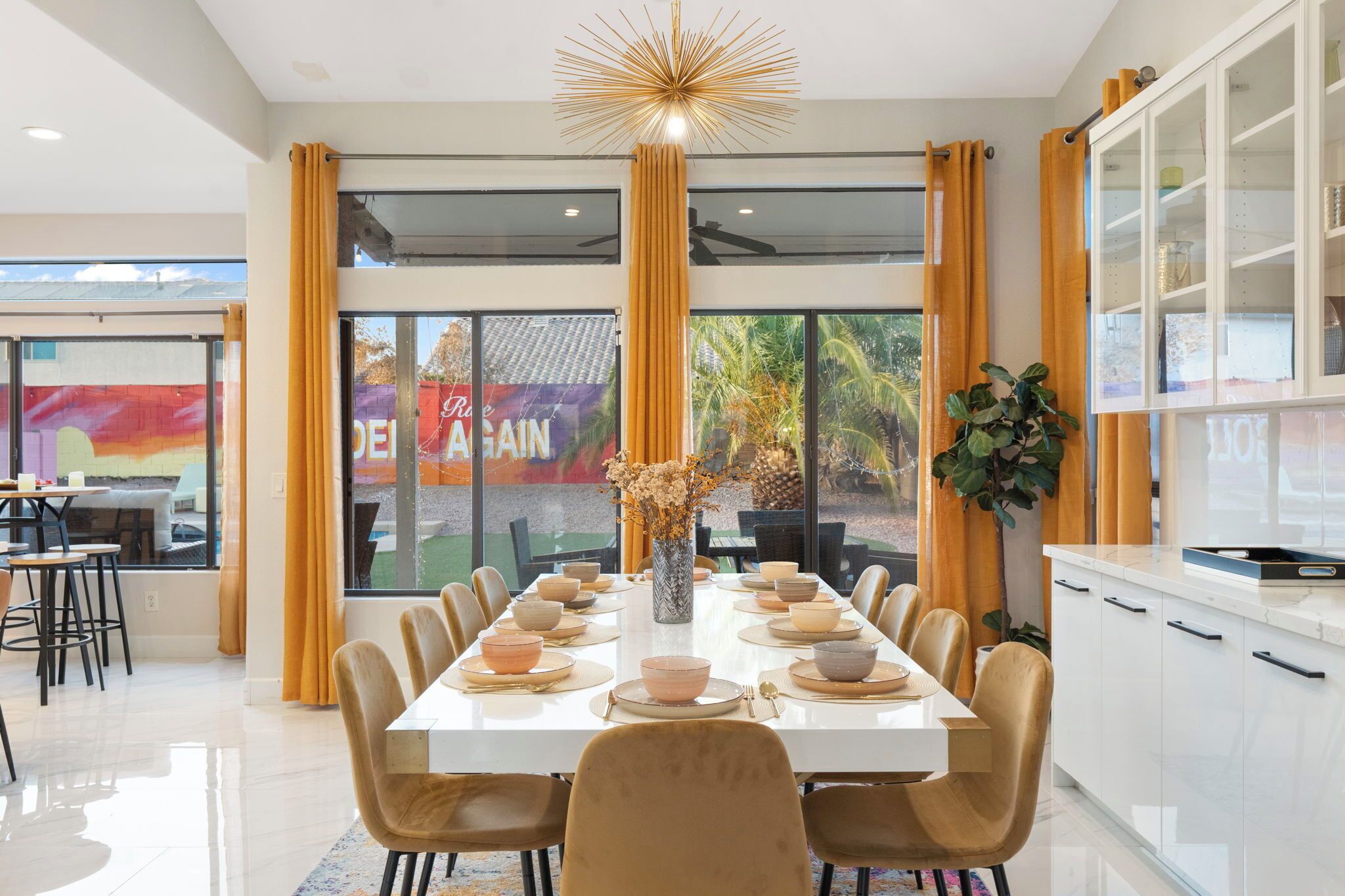 A modern dining area with a long table set for a meal, large windows with curtains, and a view of the outside.