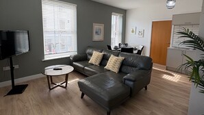 A modern living room with a leather sofa, a coffee table, and a TV. There are large windows with blinds, a ceiling light, and a kitchen area visible in the background. - 2 x bed city apartment with parking  (Hull)