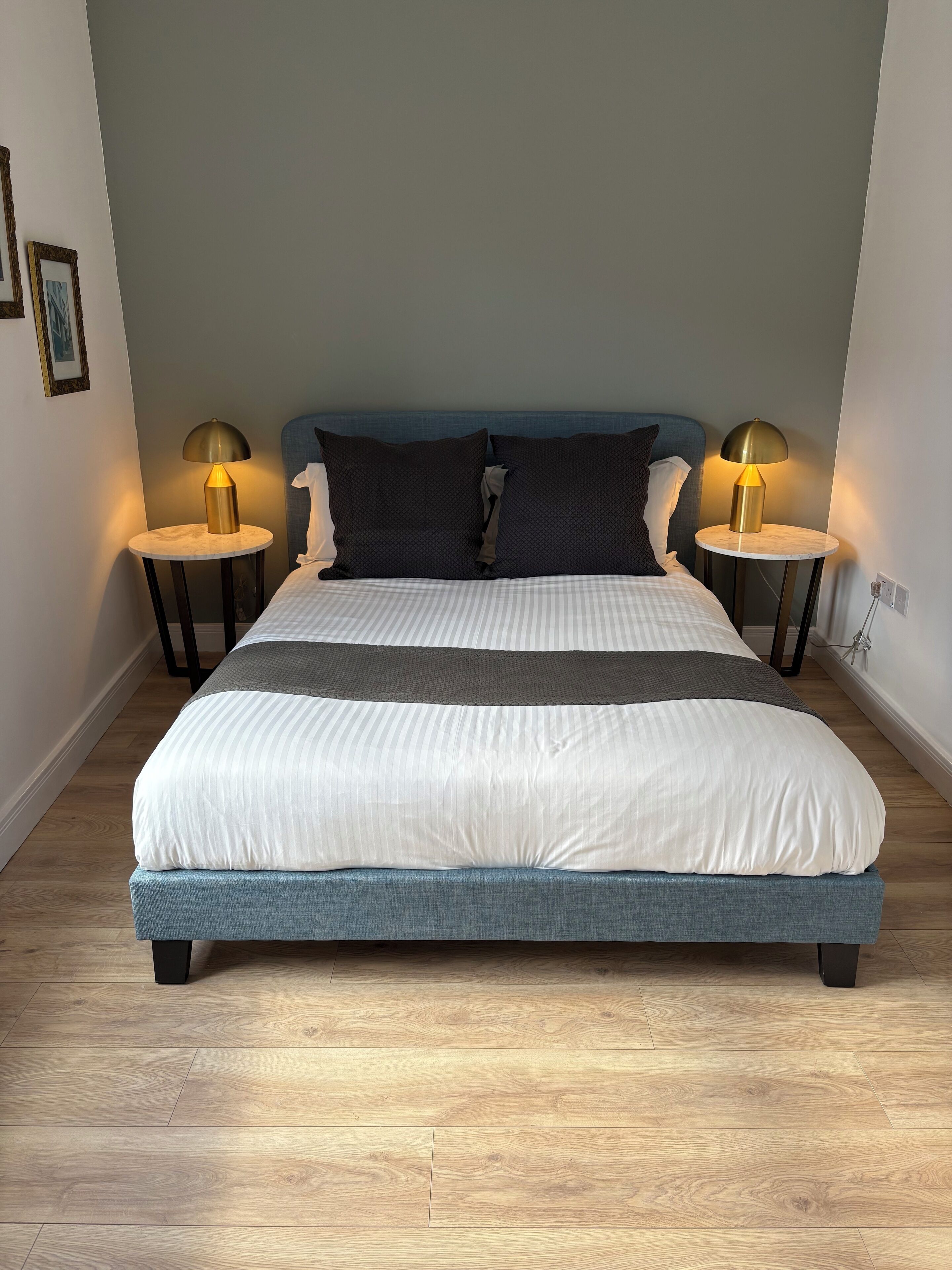 Een netjes opgemaakt bed met een blauw hoofdbord, twee bedlampjes en een houten vloer.