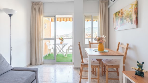 Un salon moderne avec un canapé gris, un coin repas avec une table et des chaises en bois, et un balcon offrant une vue sur la ville.