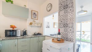 Une cuisine compacte équipée d’un micro-ondes, d’un grille-pain et d’une machine à café sur le plan de travail. On y trouve également un réfrigérateur avec un panier sur le dessus. La cuisine est dotée de meubles de couleur vert clair et d’un crédence à motifs. Une horloge et un tableau encadré ornent le mur.