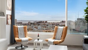Habitación moderna con dos sillones, una mesita con una botella y un ventanal con vistas a un paisaje urbano.