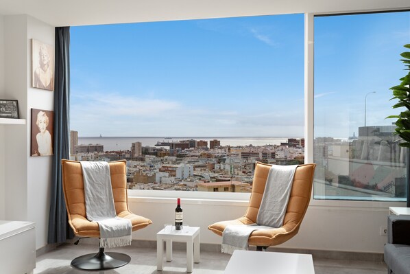 Habitación moderna con dos sillones, una mesita con una botella y un ventanal con vistas a un paisaje urbano.