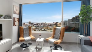 Una sala moderna con dos sillones de cuero, una pequeña mesa blanca con una botella, y un ventanal que ofrece vistas de la ciudad.