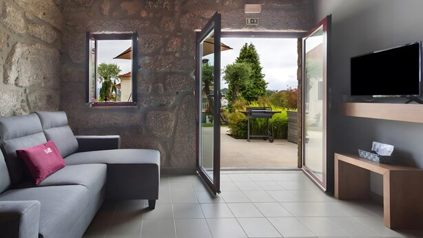 A modern room with a stone wall, a grey sofa with a pink pillow, a television, and a sliding glass door leading to an outdoor area. - Holiday Home 'Lagar T1' with Lake View, Wi-Fi & Air Conditioning (Vieira do Minho)