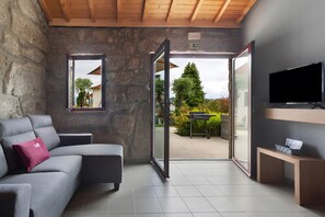 Une chambre moderne avec un mur en pierre, un canapé gris agrémenté d’un coussin rose, une télévision et une porte-fenêtre coulissante donnant sur un espace extérieur.