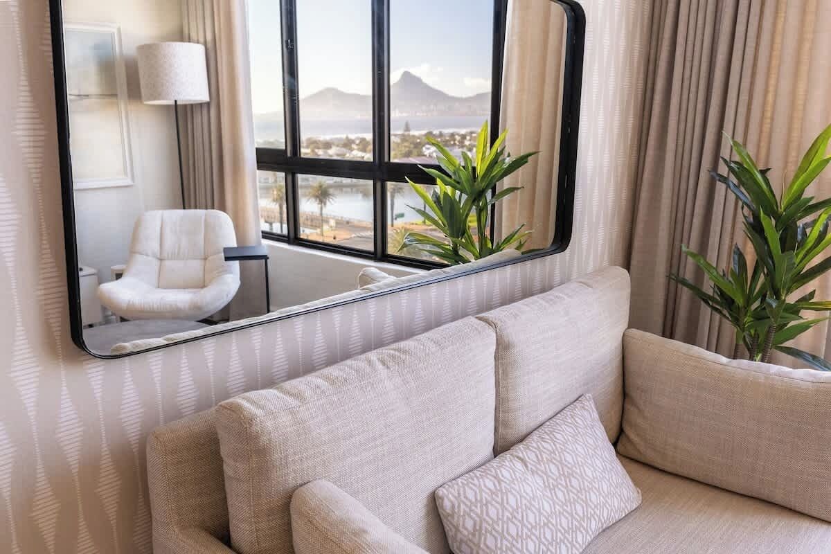 Un salon moderne avec un canapé, un fauteuil, un grand miroir reflétant un paysage et une plante en pot.