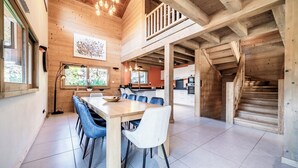 Ein großzügiger Essbereich mit einem langen Holztisch und blauen Stühlen, eine Küche im Hintergrund und eine Treppe, die zu einer oberen Ebene führt.