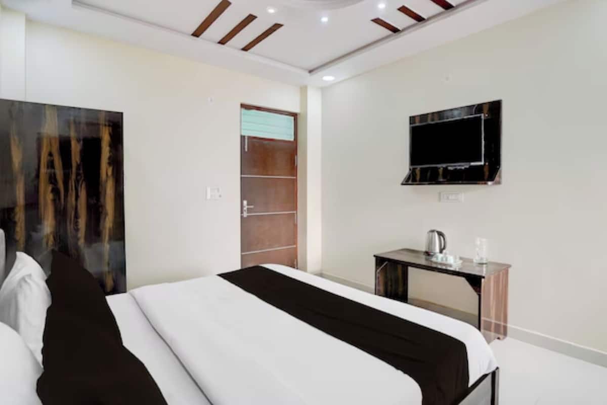 A modern hotel room with a large bed, a desk with a kettle, a flat-screen TV, and a door leading to a bathroom.