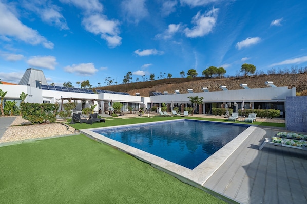 A swimming pool with a surrounding deck, a building with solar panels, and a hilly landscape with trees.