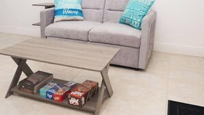 Ein Wohnzimmer mit einem grauen Sofa, zwei türkisfarbenen Kissen, einem Couchtisch aus Holz mit Gesellschaftsspielen und einem Fenster mit Jalousien.