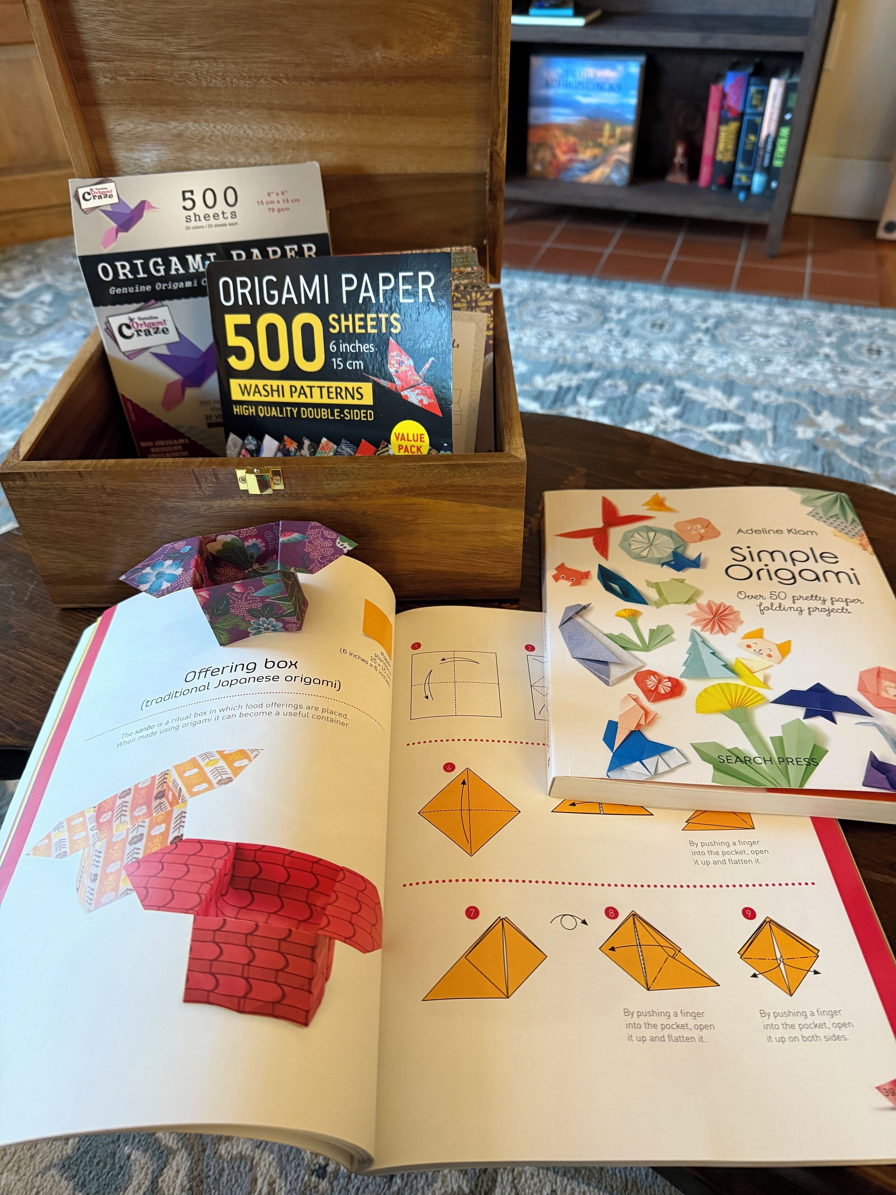 A book open on a table, a box of origami paper, and a bookshelf in the background.