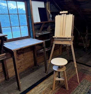 Una habitación rústica de madera con un caballete, un taburete pequeño y una ventana con vistas al agua.