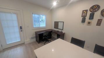 Une pièce avec un bureau blanc, un fauteuil de bureau noir et une grande fenêtre équipée de stores.