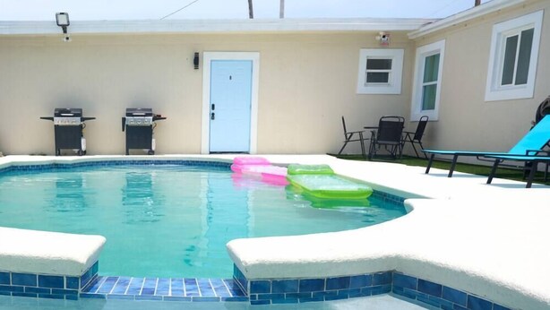 A swimming pool with a tiled edge, a door, and outdoor furniture including chairs and a table.