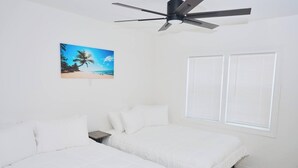 A bedroom with two beds, a ceiling fan, a window with blinds, and a painting of a beach scene.