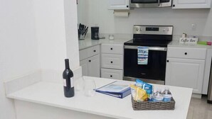 A modern kitchen with white cabinets, a microwave, a coffee maker, and a counter with a bottle, glasses, and snacks.